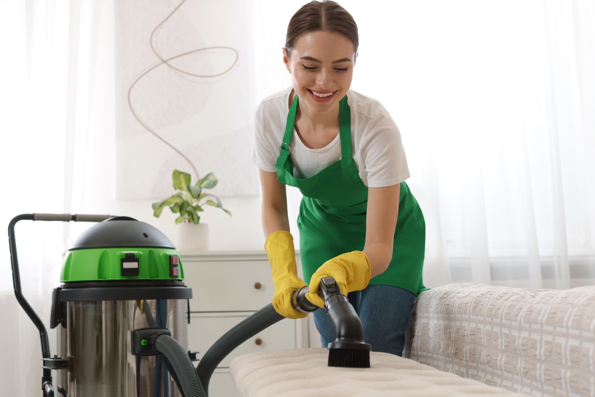 Woman in green apron and yellow gloves vacuums a sofa in a bright room.