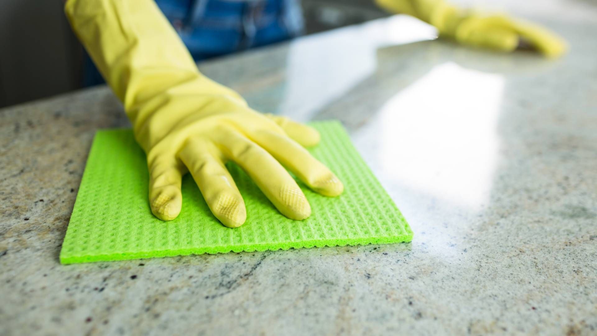 Person wearing yellow gloves wiping a light-colored countertop with a green sponge.