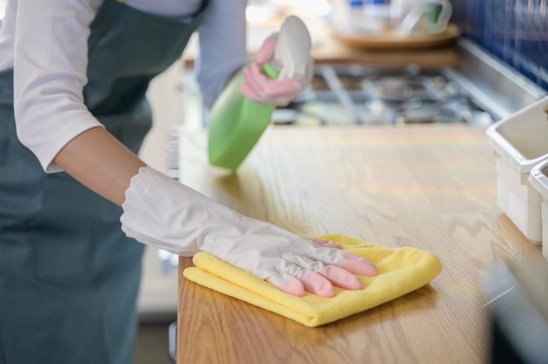 Person wearing gloves cleaning a wooden kitchen countertop with a yellow cloth and spray bottle.