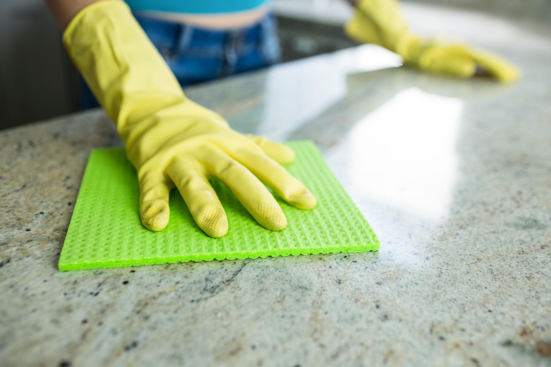Person wearing yellow gloves cleans a light-colored countertop with a green sponge.