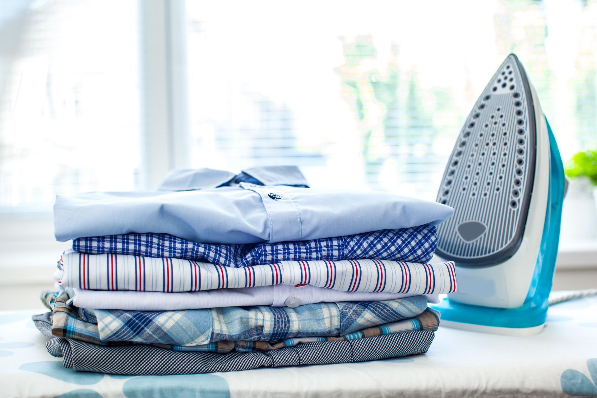 Stack of folded shirts next to an iron on an ironing board near a window.