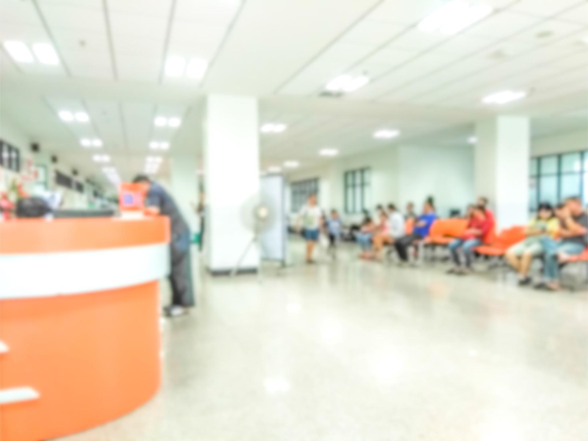 Blurred image of a bright waiting room with people seated on orange chairs and someone at a service counter.