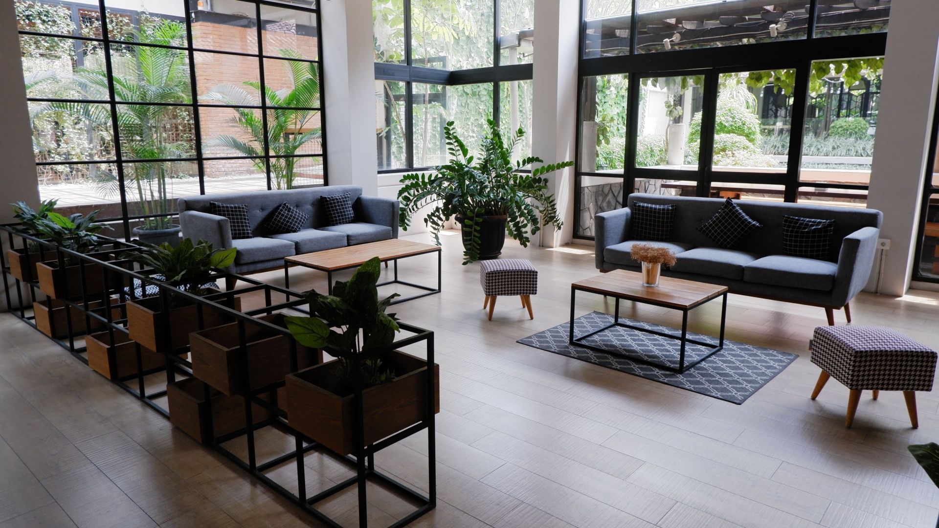 A bright, modern lobby lounge with two grey sofas, wooden coffee tables, potted plants, and floor-to-ceiling windows.