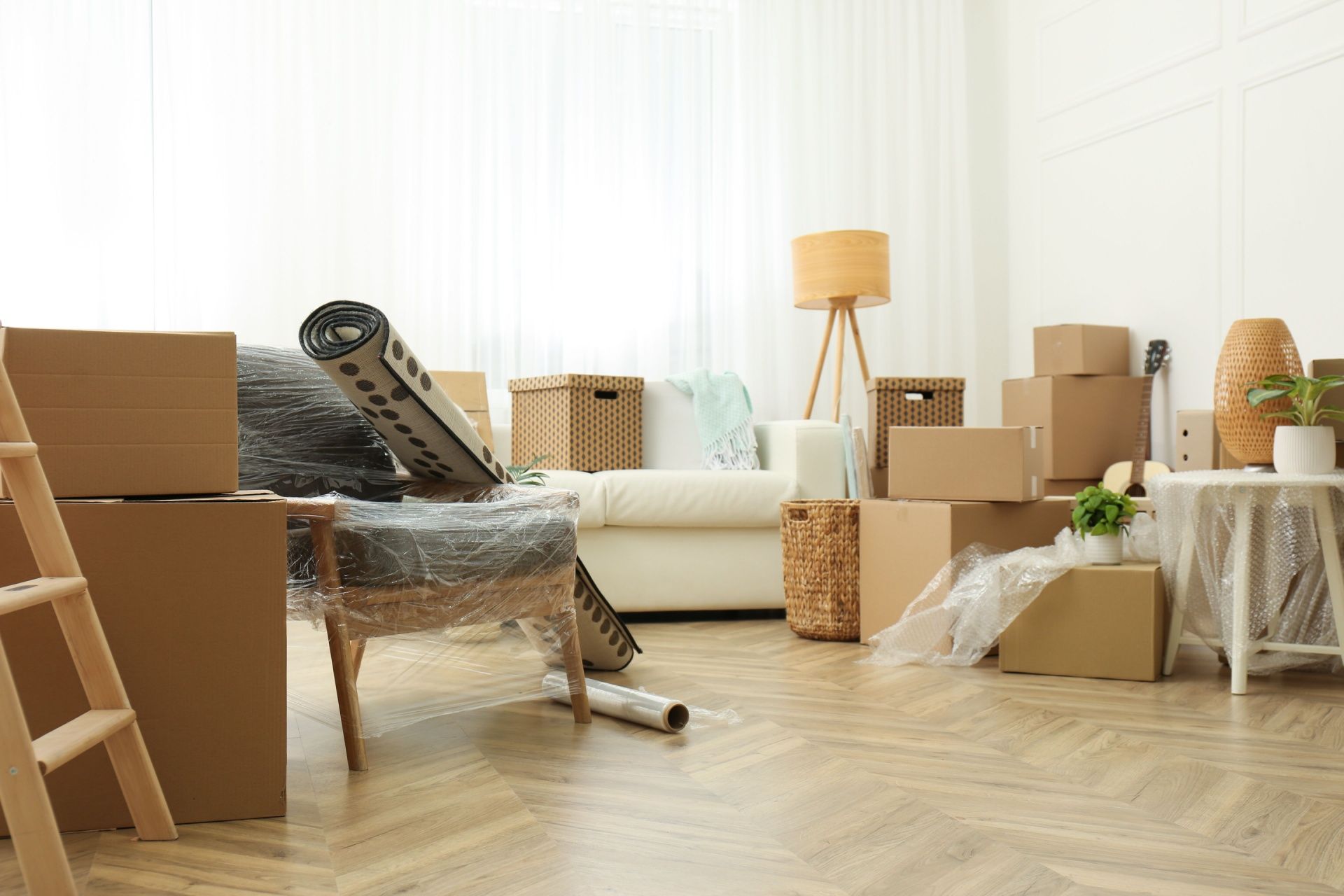 Living room filled with cardboard boxes, furniture wrapped in plastic, and a wooden ladder during a home move.