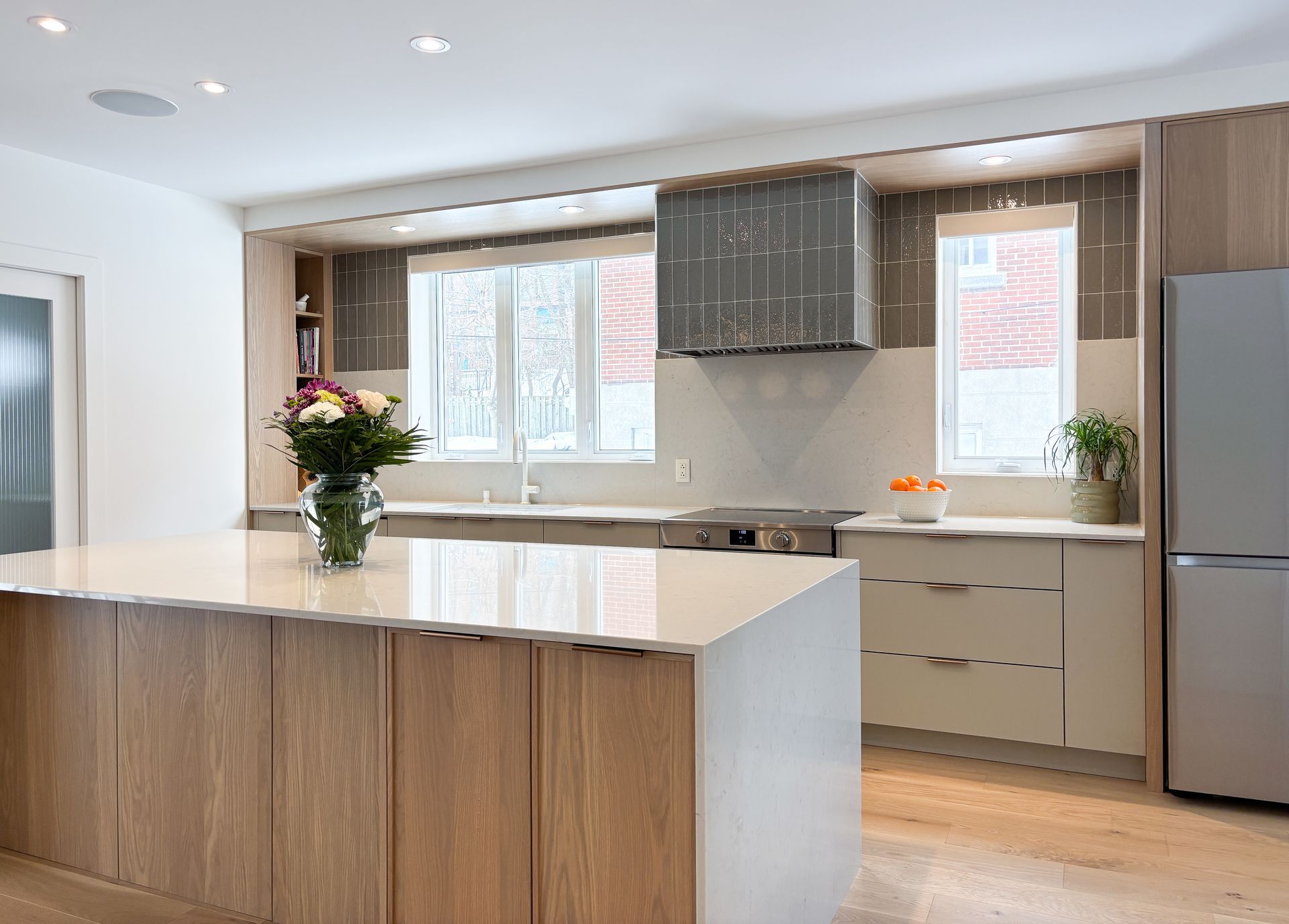 Cuisine moderne avec armoires en bois clair, comptoirs blancs et réfrigérateur en acier inoxydable.