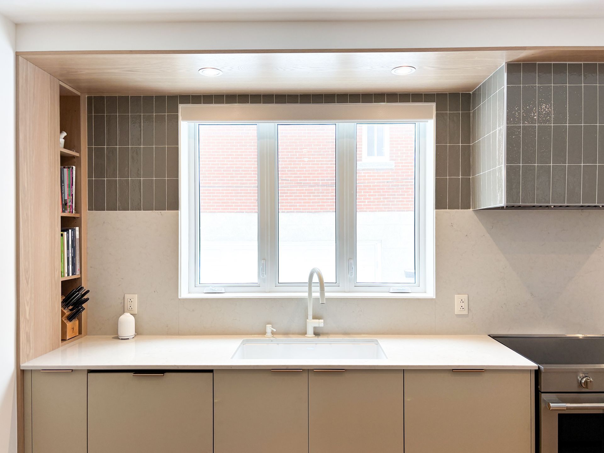 Cuisine moderne avec armoires beiges, comptoir blanc, fenêtre et dosseret carrelé gris.