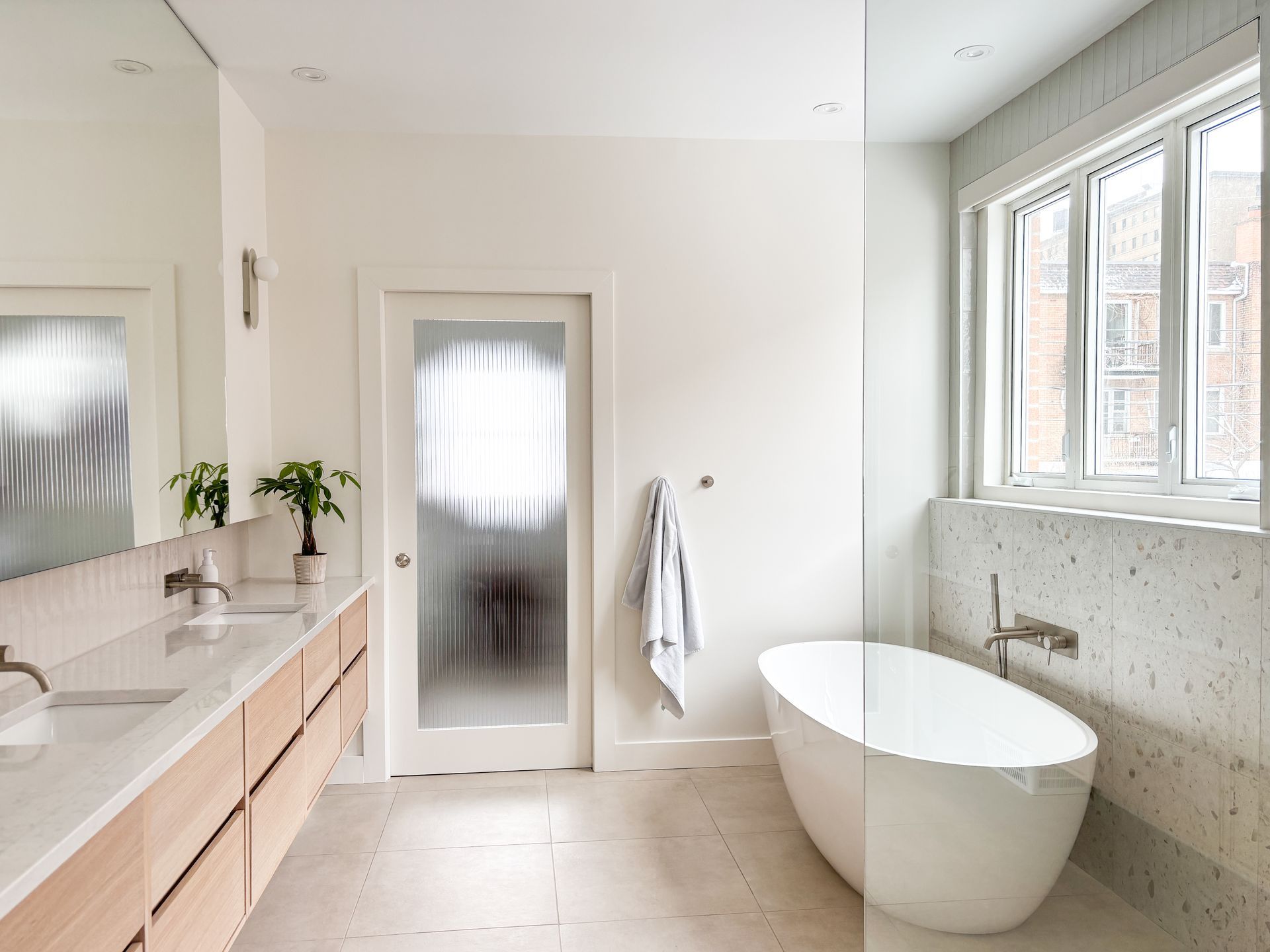 Salle de bain blanche moderne avec une baignoire autoportante, une vanité en bois clair et une porte de douche en verre texturé.