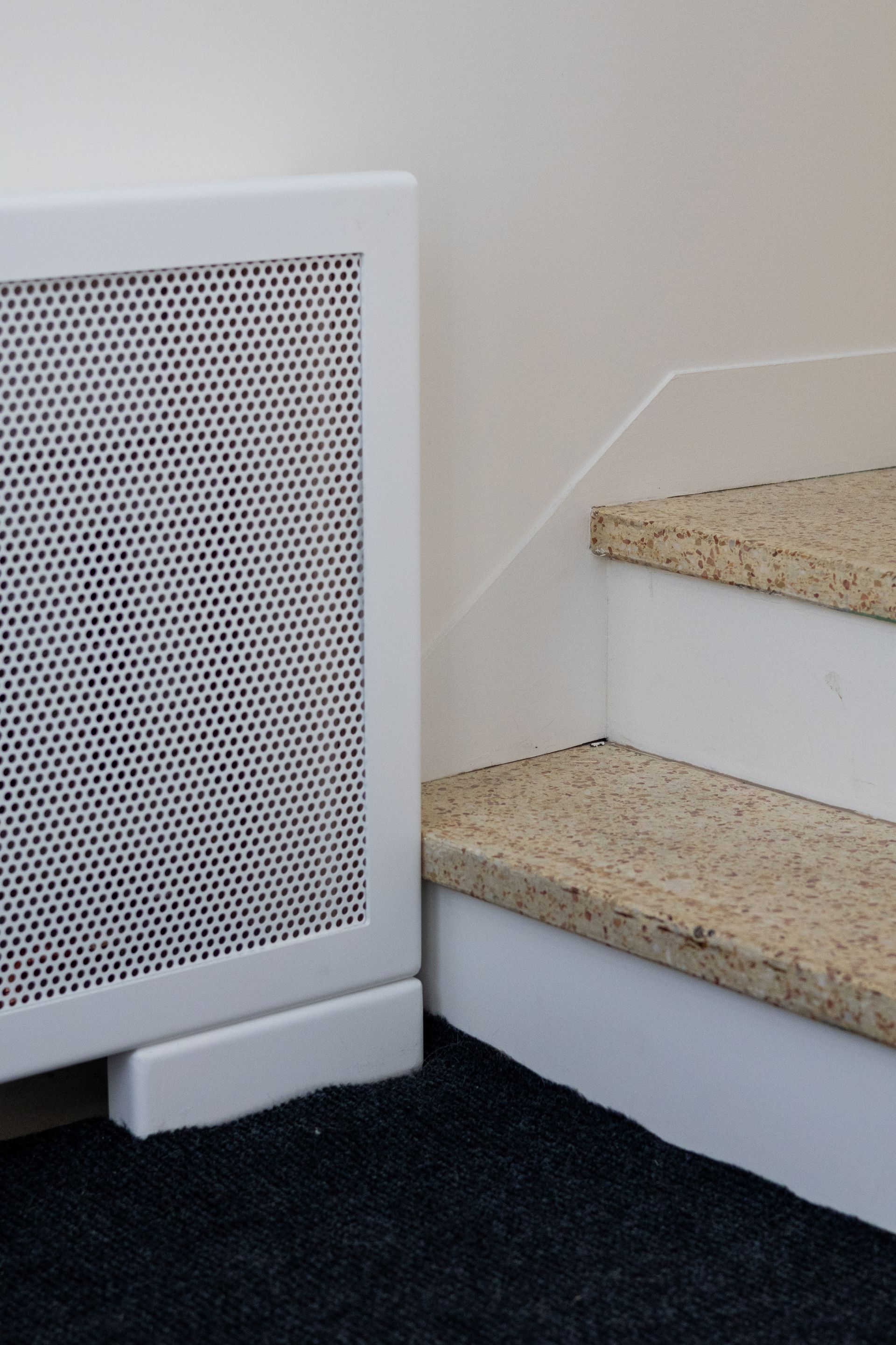 Cache radiateur blanc à côté des escaliers avec marches recouvertes de moquette beige.