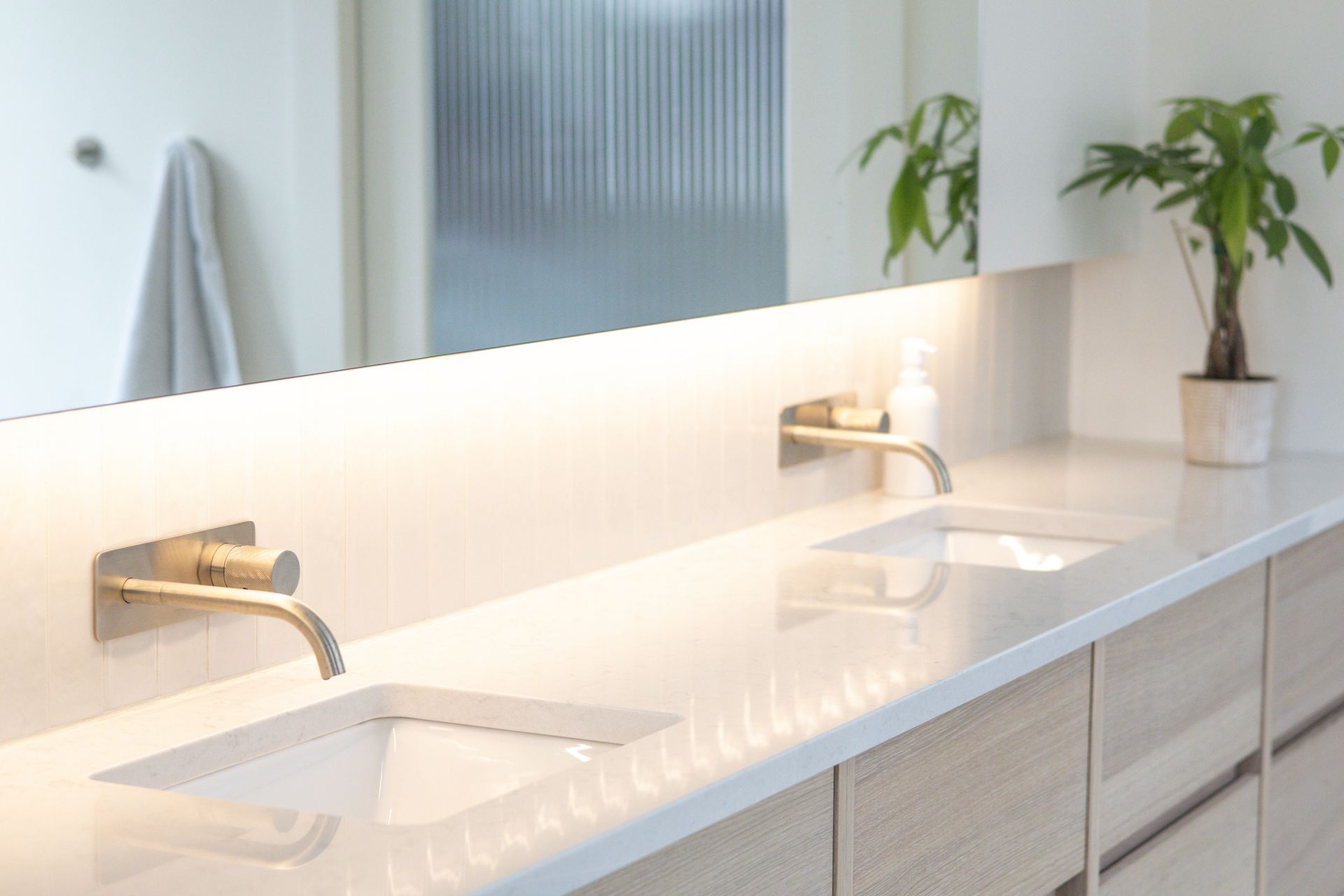Salle de bain moderne avec deux lavabos, robinets dorés, comptoir blanc, armoires en bois et une plante en pot.
