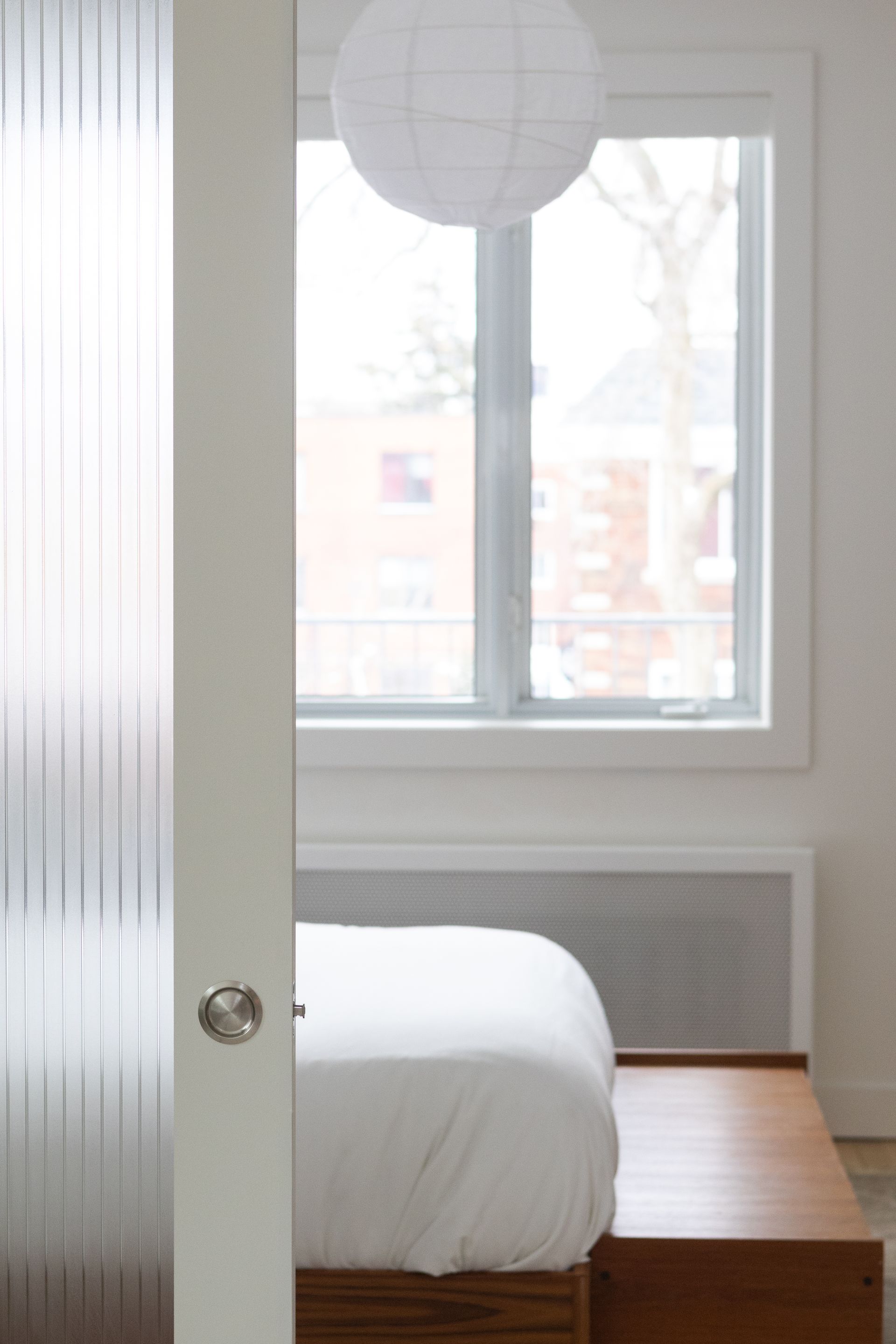 Intérieur de chambre avec un lit blanc, une fenêtre, une lanterne en papier et une porte en verre dépoli partiellement visible.