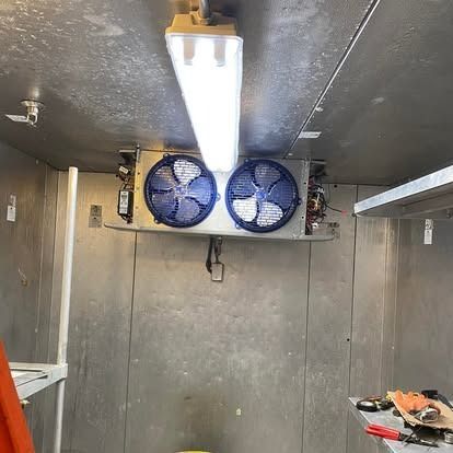 Interior of a walk-in cooler with a bright ceiling light above an industrial fan unit mounted on the metal back wall.