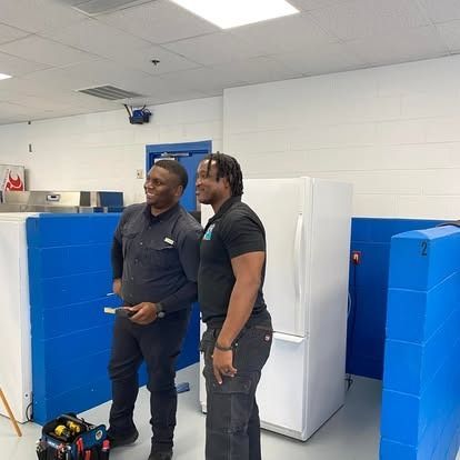 Two individuals in dark uniforms stand in a brightly lit industrial workspace with blue partitions and white freezers.