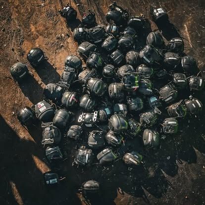 A high-angle view of many dark, modular motorcycle helmets scattered across a patch of dry, dark earth.