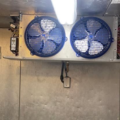 A commercial refrigeration unit mounted on a wall with two blue circular fan guards and a digital thermostat controller.