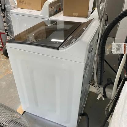 A high-angle view of a white top-loading washing machine with a dark glass lid, sitting in an indoor storage area.