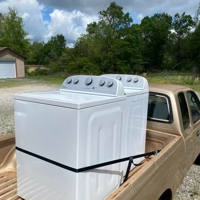 Two white washing machines secured with a black strap in the bed of a tan pickup truck against a backdrop of trees.