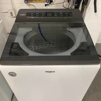 A white Whirlpool top-load washing machine with a dark glass lid, situated in a utility area.
