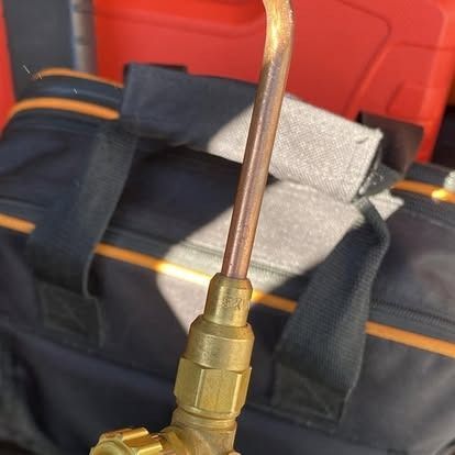 A brass torch head with a curved metal tube attached, positioned in front of a dark gear bag and a red case.