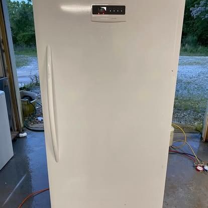 A white upright freezer stands in an outdoor area, featuring a tall vertical handle and a digital control panel on top.