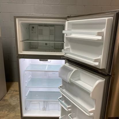 An open, empty, stainless steel top-freezer refrigerator against a gray concrete block wall.