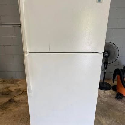A plain, white, top-freezer refrigerator stands in an indoor space with a fan visible in the background.