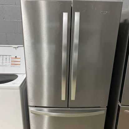 A stainless steel Whirlpool French-door refrigerator with a bottom freezer drawer, positioned next to a washing machine.