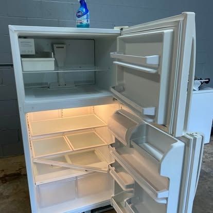 An open, white, empty top-freezer refrigerator stands in a room with gray cinder block walls.