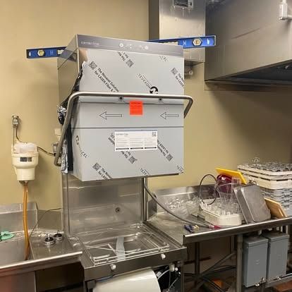 A stainless steel commercial dishwasher in a kitchen, topped with a blue spirit level, surrounded by workspace equipment.