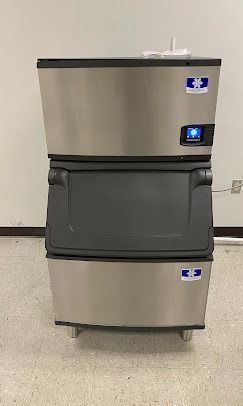 A stainless steel Manitowoc ice machine with a dark gray central bin, standing on a light-colored floor against a wall.