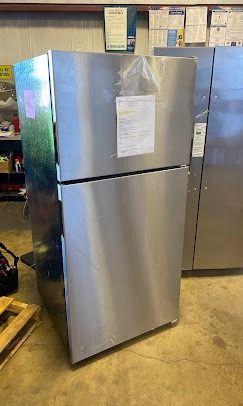 A stainless steel top-freezer refrigerator stands on a concrete floor in a warehouse setting.