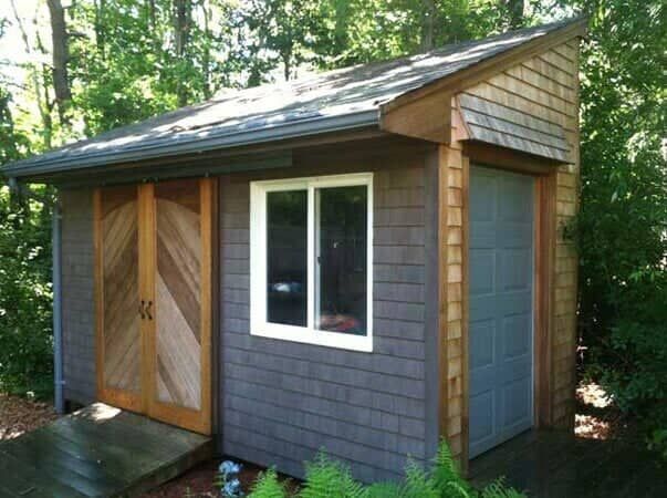 A shed with brown shingle siding, a single window, wooden double doors, and a separate side door, set in a wooded area.
