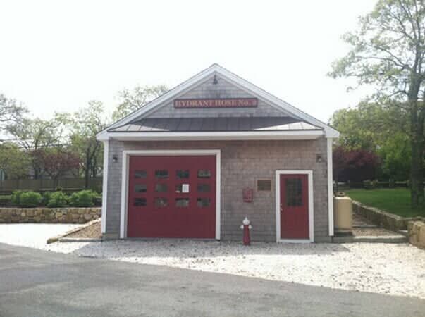 A small, shingled fire station building with a large red garage door, a red side door, and a sign reading 