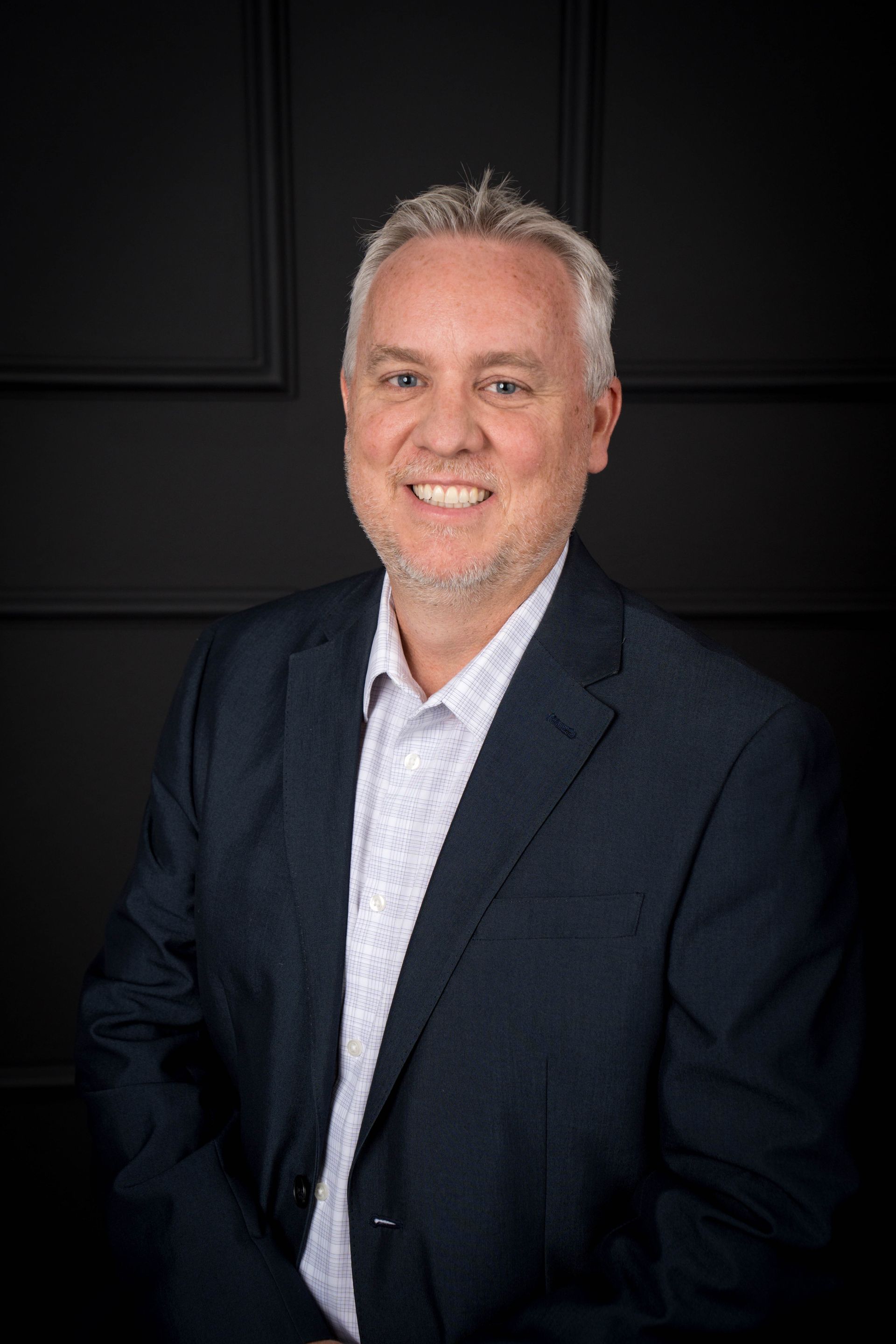 A smiling person in a blue suit and light patterned dress shirt, positioned against a dark, paneled wall.