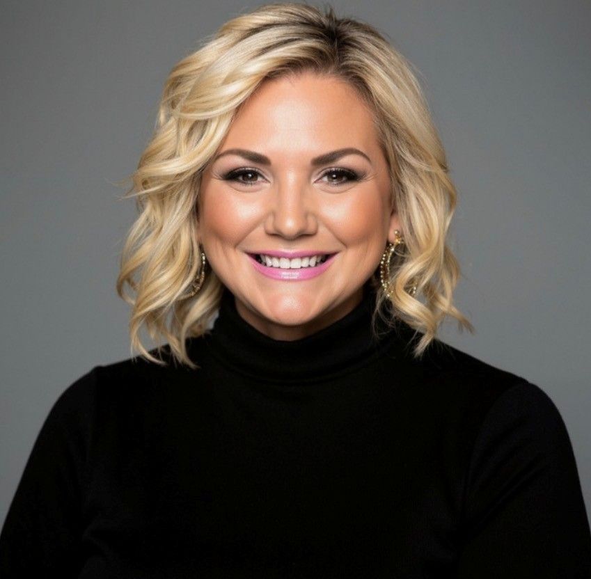 A smiling person with shoulder-length blonde hair, wearing a black turtleneck, posed against a solid grey background.