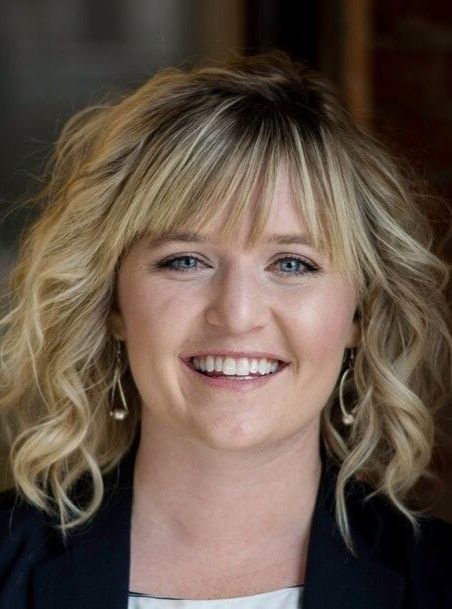 A smiling woman with shoulder-length wavy blonde hair and bangs, wearing a dark blazer, shown in a professional headshot.