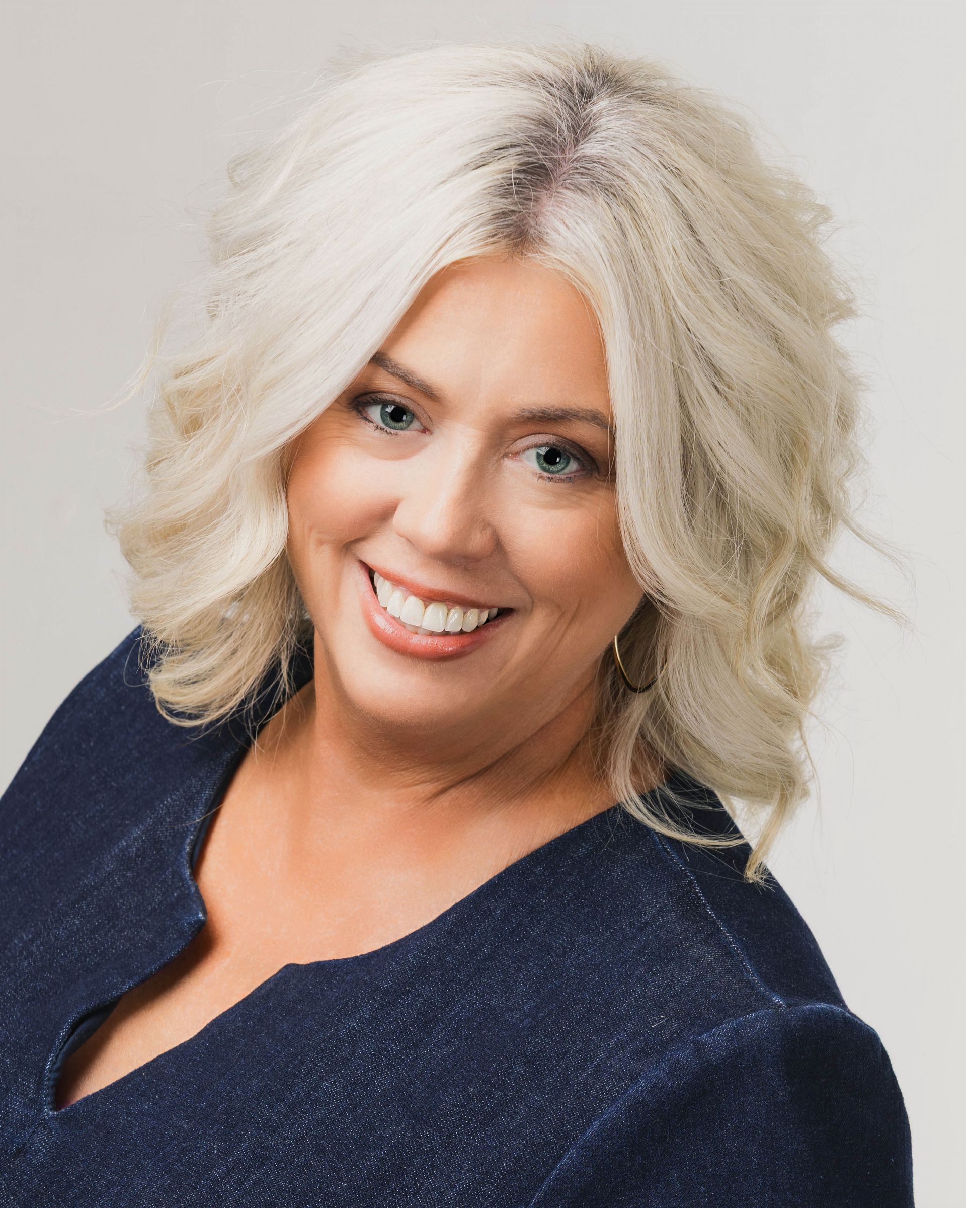 A smiling woman with shoulder-length, layered blonde hair, wearing a dark blue textured top against a light gray background.