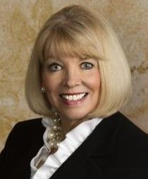 A blonde woman wearing a black jacket and white ruffled shirt smiling against a tan background.