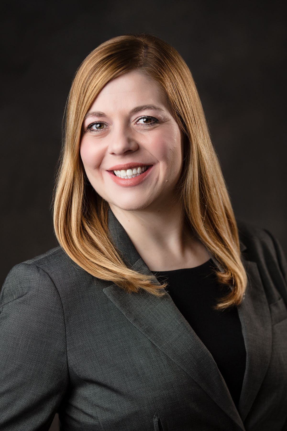 A person with shoulder-length blonde hair wearing a grey blazer over a black top, smiling against a dark background.