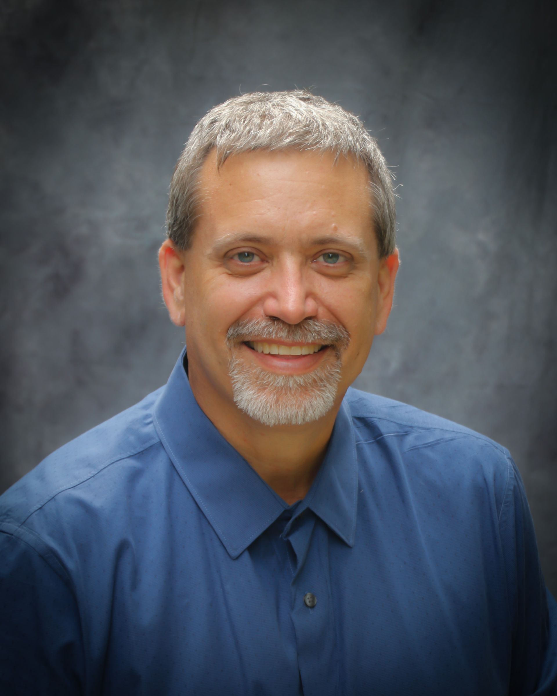 A person with short, graying hair and a goatee smiles at the camera, wearing a blue collared shirt against a gray backdrop.