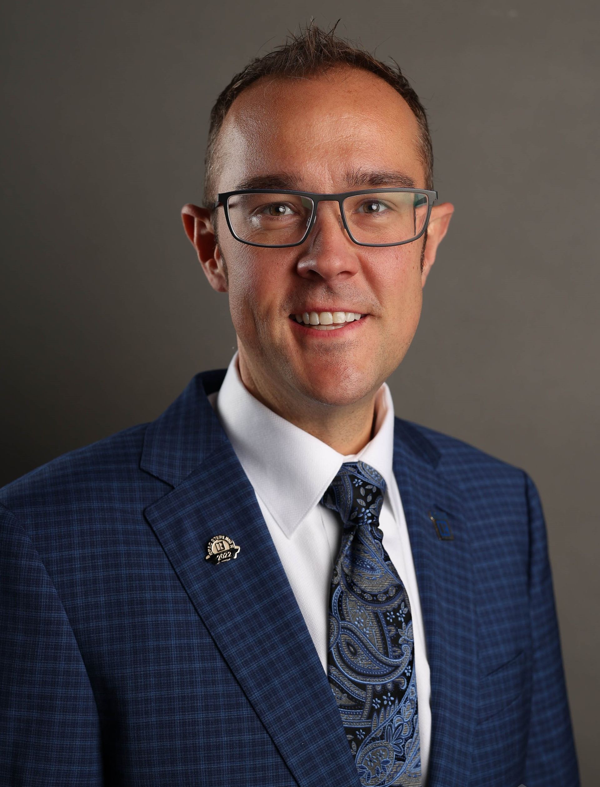 Professional headshot of a person wearing glasses, a blue patterned blazer, a white collared shirt, and a paisley tie.