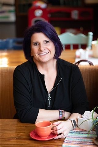 A person with purple hair wearing a black top sits at a wooden table, smiling while holding an orange cup and saucer.