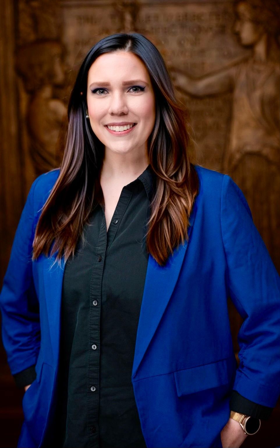 A person with long dark hair, wearing a blue blazer over a black button-down shirt, smiles in front of a textured wall.