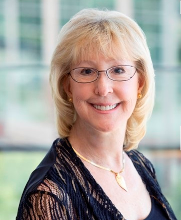Headshot of a person with blonde hair and glasses, smiling, wearing a black lace top and gold pendant necklace.
