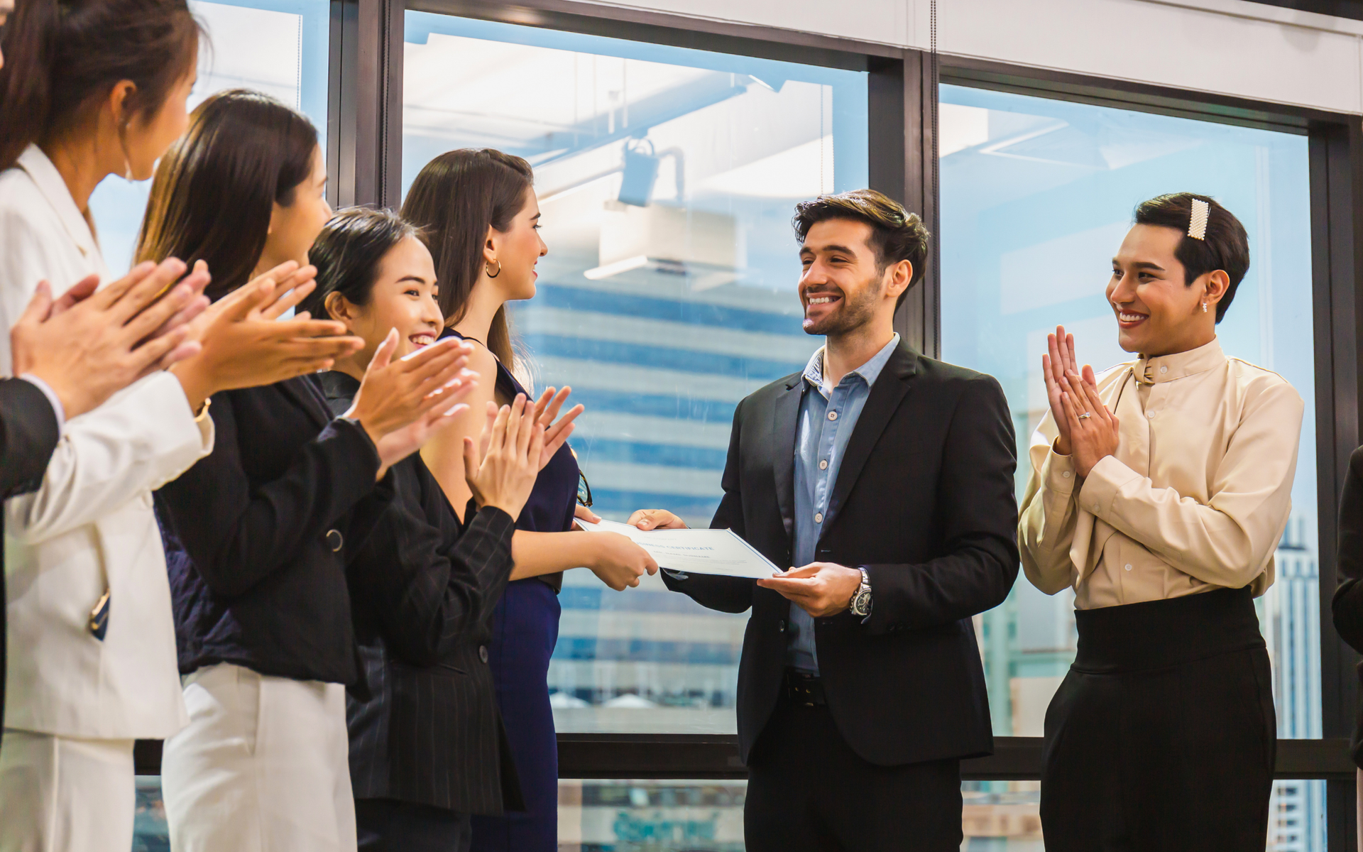A group of professionals in an office clapping as a person in a suit receives a document.