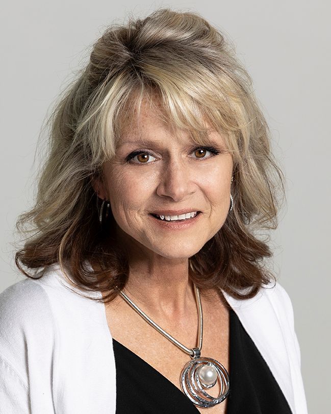 A person with blonde-highlighted hair smiling in a black top and white cardigan, wearing a circular silver pendant necklace.