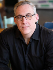 A person with short grey hair and glasses, wearing a black button-down shirt, looking directly into the camera.