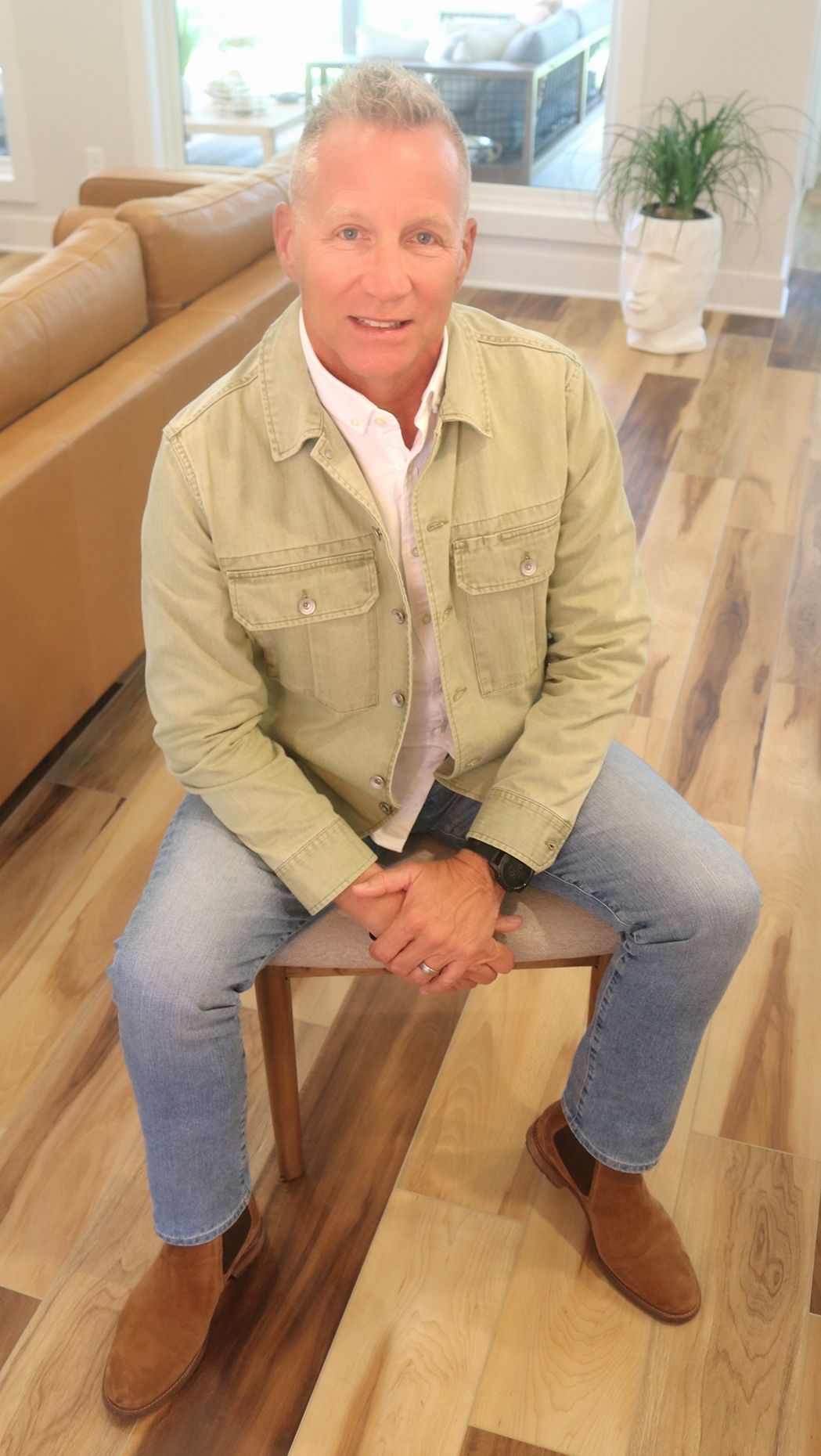 A person wearing a light green jacket and blue jeans sitting on a wooden chair in a modern, light-filled living space.