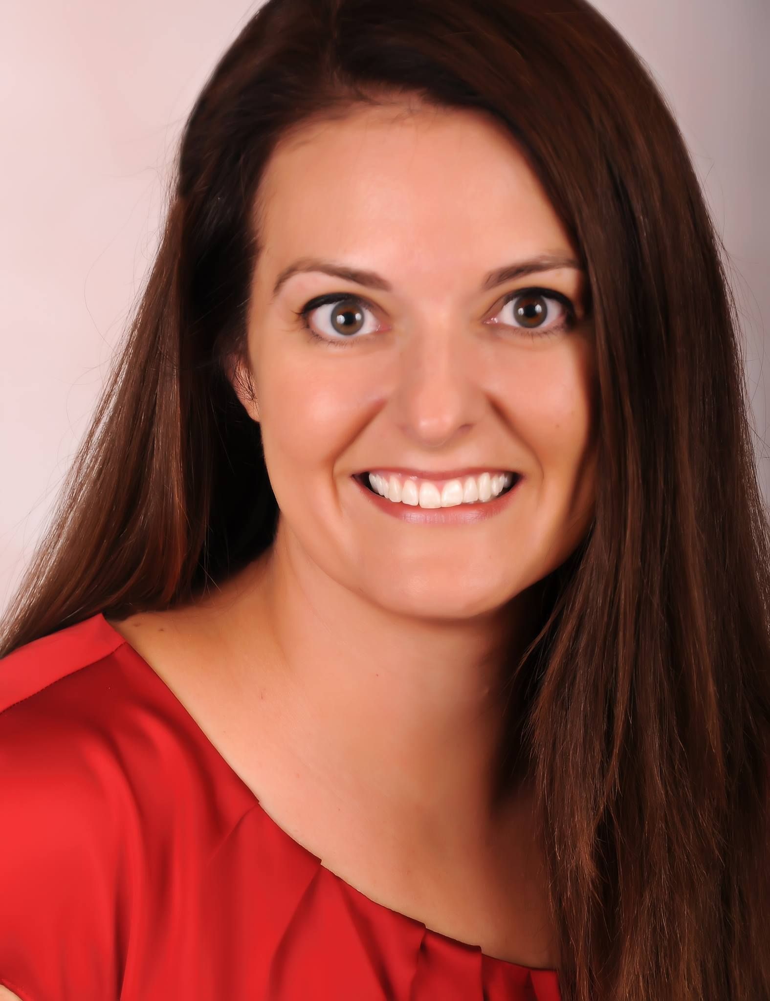 A person with long brown hair smiling at the camera, wearing a red top against a neutral background.