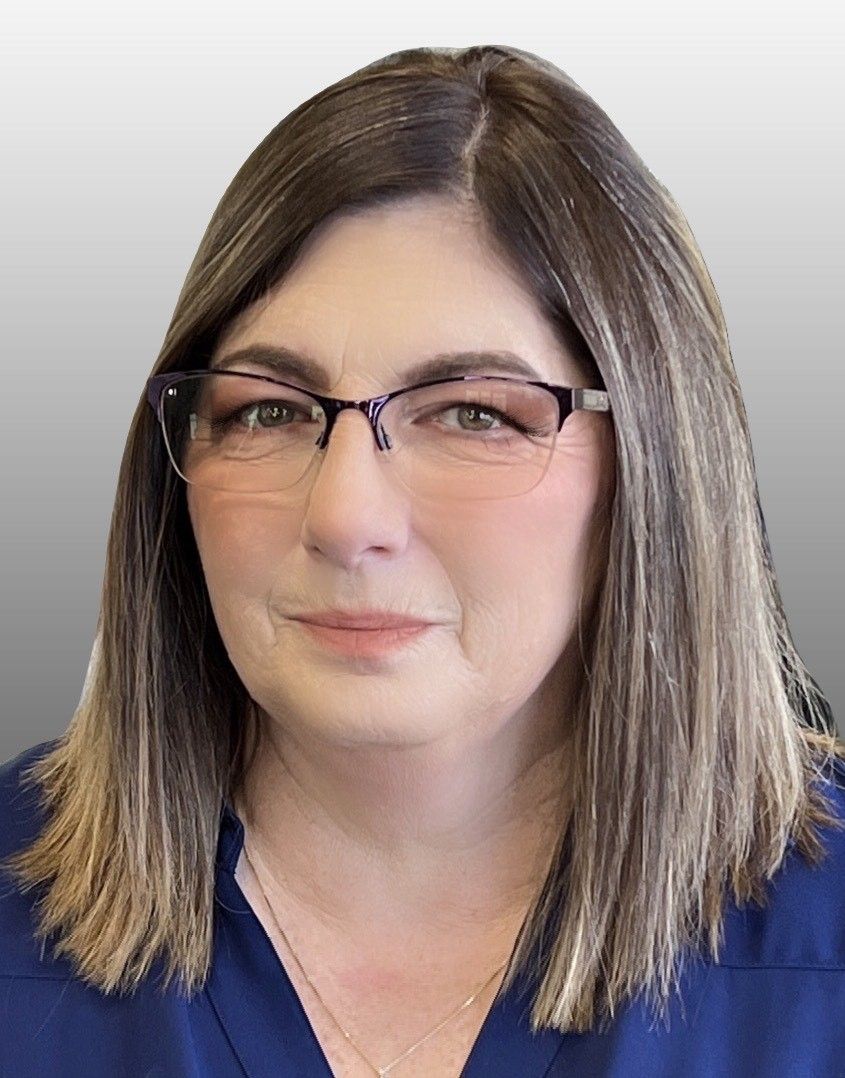 A person with shoulder-length brown hair and glasses wearing a blue top against a neutral gray background.