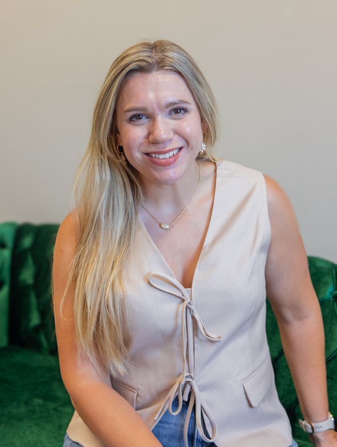 A smiling person with long blonde hair wears a beige sleeveless top and necklace, sitting on a dark green tufted sofa.